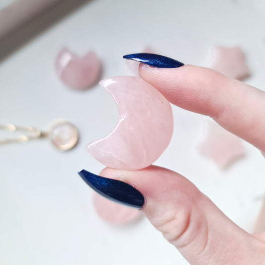 A photo of a moon carving made from rose quartz, a type of quartz crystal with a soft pink colour. The surface of the moon is polished to a smooth, shiny finish, with natural variations in texture and colour visible in the stone.