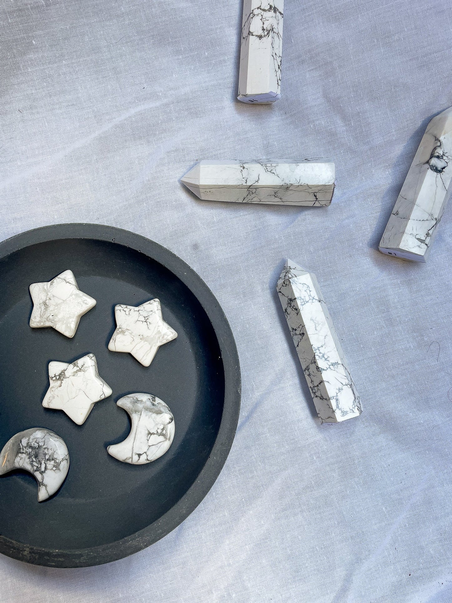 A photo of howlite moon and star carvings arranged in a grey bowl, along with several howlite points beside the bowl, against a light grey background.